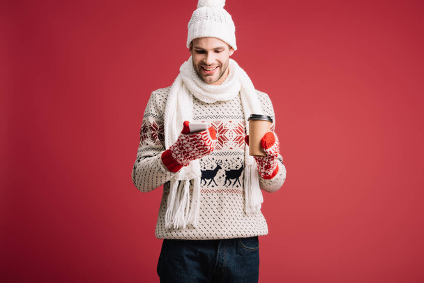 smiling man in winter sweater, scarf, hat and mittens holding coffee to go while using smartphone, isolated on red - Foto, immagini