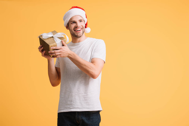 cheerful man in santa hat holding christmas present isolated on yellow - Foto, imagen