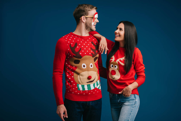 smiling man in glasses with santa hat hugging girlfriend isolated on blue - Фото, изображение