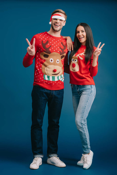 boyfriend in glasses with santa hat and cheerful girlfriend showing victory signs isolated on blue - Foto, Imagem