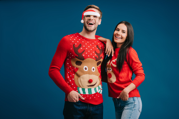 boyfriend in glasses with santa hat hugging cheerful girlfriend isolated on blue - Foto, imagen