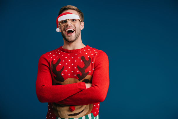 Aufgeregter Mann posiert in Weihnachtspullover und Brille mit Weihnachtsmütze, isoliert auf blauem Grund - Foto, Bild