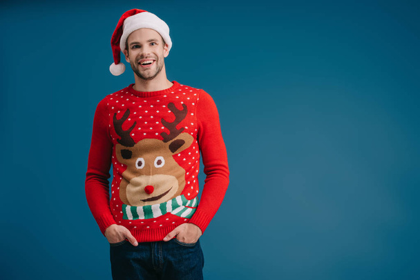 smiling man posing in santa hat and christmas sweater isolated on blue - 写真・画像