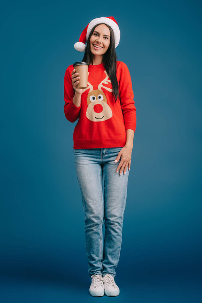 smiling girl in santa hat and christmas sweater holding coffee to go, isolated on blue - Fotoğraf, Görsel