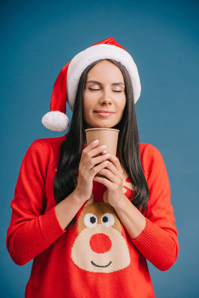 attractive woman with closed eyes in santa hat and christmas sweater holding coffee to go, isolated on blue - Foto, Bild