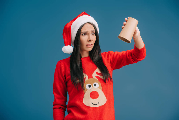 upset woman in santa hat holding empty cup of coffee, isolated on blue - Foto, afbeelding
