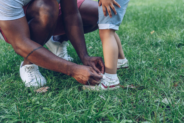 cropped view of african american man tying shoelaces of son in park - Zdjęcie, obraz