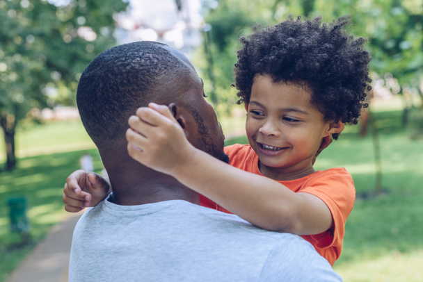 cute african american boy hugging father while walking in park - 写真・画像