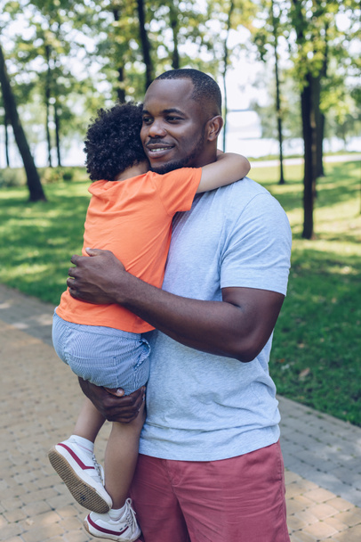 feliz afro-americano homem segurando as mãos e abraçando adorável filho no parque
 - Foto, Imagem