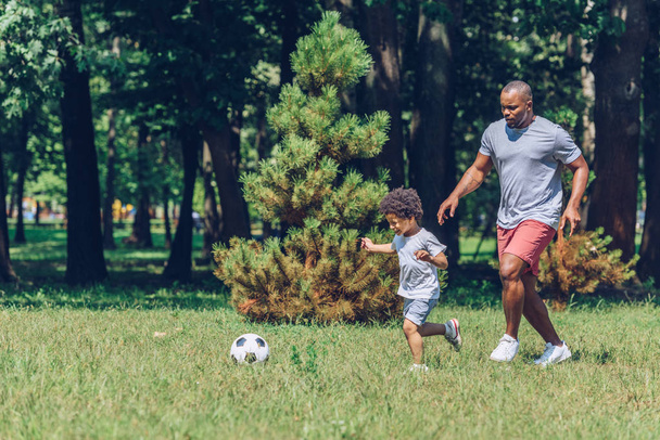 cheerful african american father and son playing football in park - Foto, afbeelding
