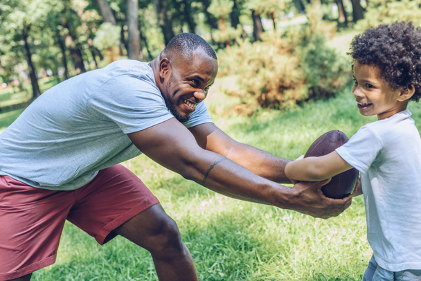 Χαρούμενος Αφροαμερικάνος που παίζει ράγκμπι με τον αξιολάτρευτο γιο του στο πάρκο - Φωτογραφία, εικόνα