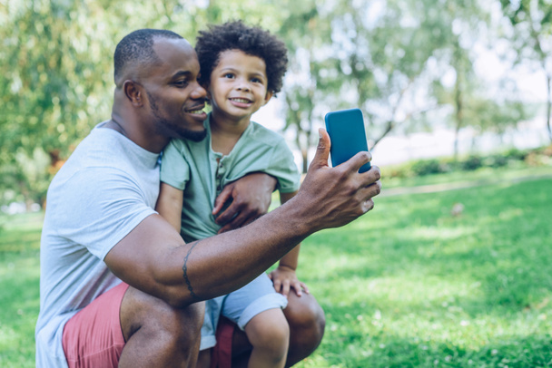cheerful african american father and son taking selfie on smartphone in park - Foto, imagen