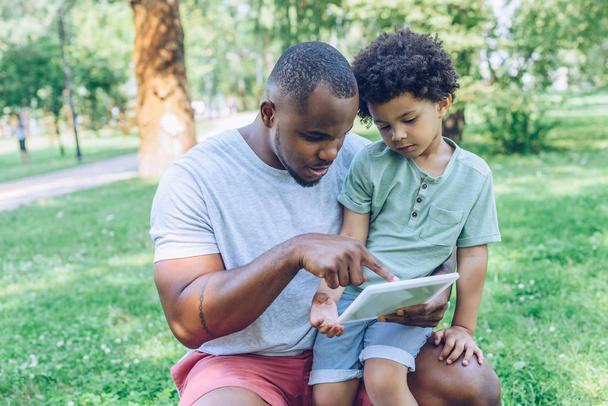 jong Afrikaans amerikaans vader met behulp van digitale tablet naar schattige zoon in park   - Foto, afbeelding