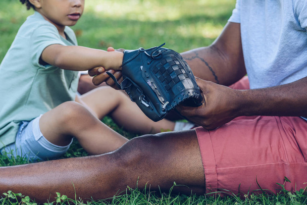 vue recadrée d'un homme afro-américain montrant un gant de baseball à son fils assis sur une pelouse dans un parc
 - Photo, image