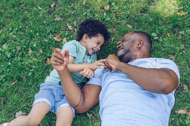 happy african american father and son having fun while lying on lawn in park - Photo, Image