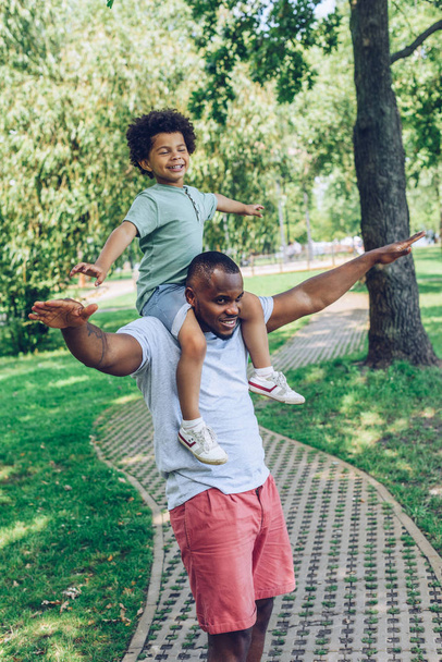 Веселый африканский американец, имитирующий полет во время игры на спине с отцом в парке
 - Фото, изображение
