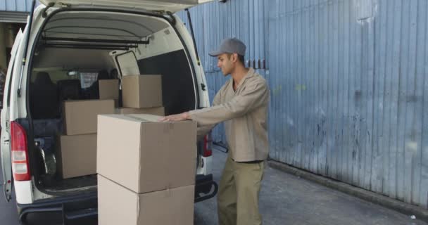 Side view of a young mixed race male warehouse worker loading boxes from a trolley into the back of an open van outside a warehouse - Imágenes, Vídeo