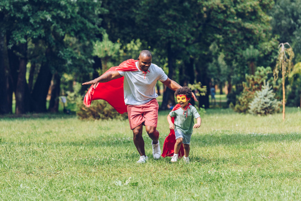 onnellinen afrikkalainen amerikkalainen isä ja poika käynnissä puvuissa supersankareita puistossa
 - Valokuva, kuva