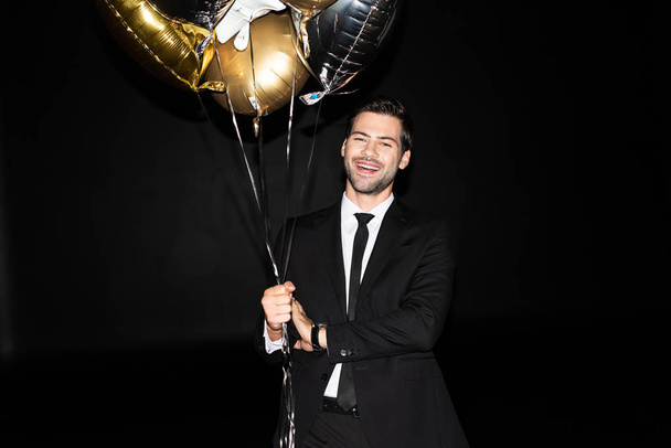 χαμογελαστός άνδρας κρατώντας χρυσά μπαλόνια για τα γενέθλια, απομονωμένος σε μαύρο - Φωτογραφία, εικόνα