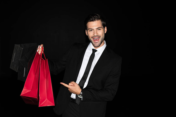 cheerful elegant man in suit pointing at shopping bags isolated on black - Photo, Image