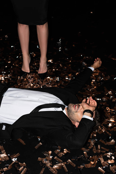 young man lying on golden confetti while girlfriend standing near on black - Zdjęcie, obraz