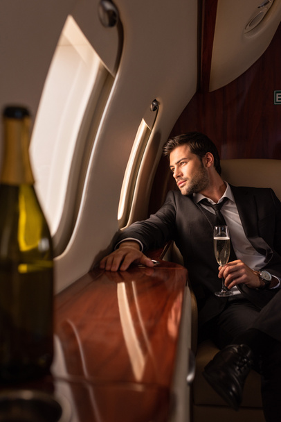 ονειροπόλος άνθρωπος κρατώντας ένα ποτήρι σαμπάνια στο αεροπλάνο, επιλεκτική εστίαση του μπουκαλιού  - Φωτογραφία, εικόνα