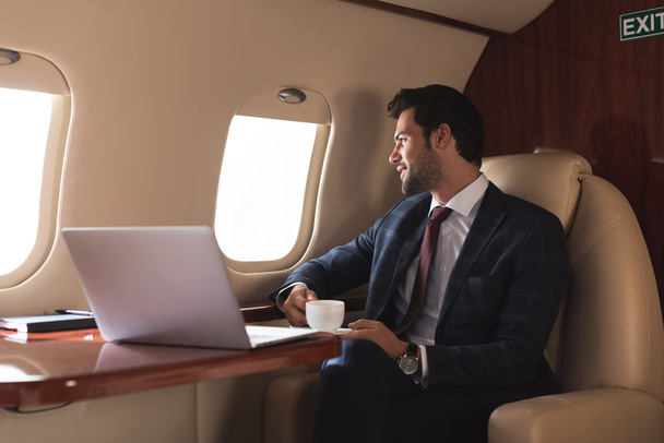 pensive businessman holding cup of coffee in plane with laptop during business trip  - Fotografie, Obrázek