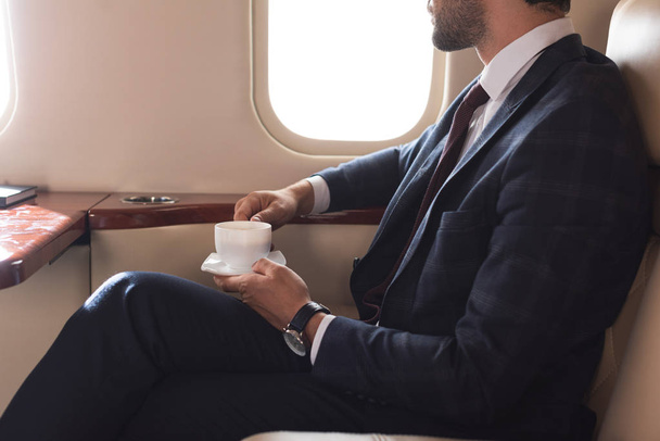 cropped view of businessman in suit holding cup of coffee in airplane during business trip - Foto, afbeelding