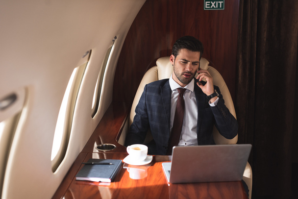 Männliche Führungspersönlichkeit spricht während Geschäftsreise im Flugzeug mit Laptop auf Smartphone  - Foto, Bild