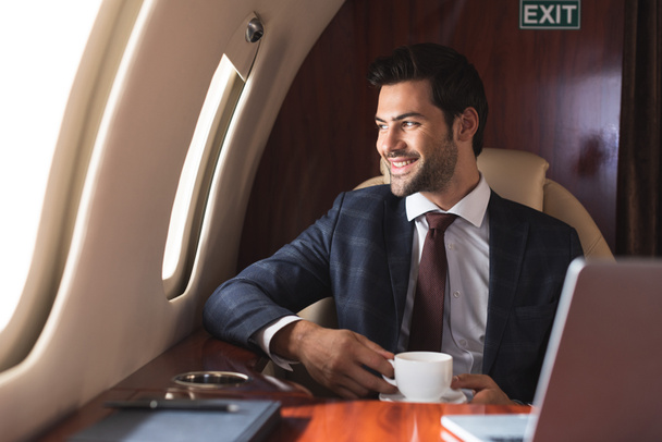 smiling businessman holding cup of coffee in plane during business trip  - Photo, Image