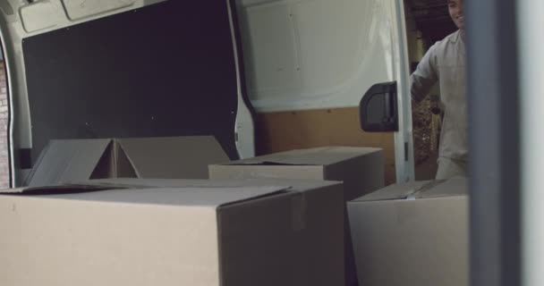 Front view of a young mixed race man opening the side door of a van full of boxes, lifting one up and carrying it into a warehouse, seen from inside the van - Materiaali, video