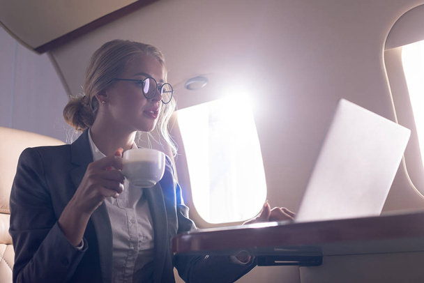 schöne Geschäftsfrau hält Kaffeetasse in der Hand und arbeitet während einer Geschäftsreise am Laptop im Flugzeug  - Foto, Bild