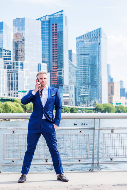 Nuori amerikkalainen liikemies, jolla on parta, puhuu kännykällä, matkustaa New Yorkissa, pukeutuu siniseen pukuun, valkoiseen paitaan, seisoo bisnesalueella, jossa on korkeat rakennukset Manhattanin keskustassa.
 - Valokuva, kuva