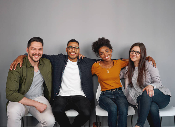 Portrait of multiethnic young friends sitting together happily with their arms around each other on chair smiling and looking at camera - Foto, immagini