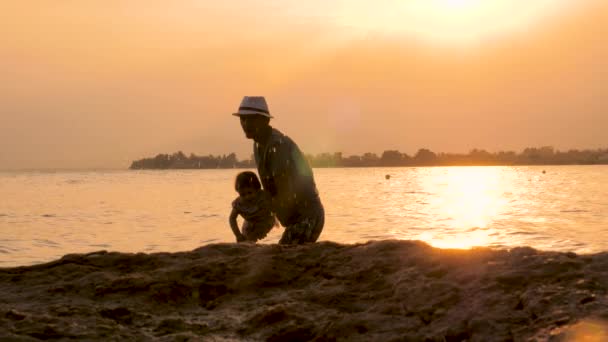 Młody ojciec i jego dziecko bawią się podczas zachodu słońca na plaży, sylwetki taty bawią się z dzieckiem w morzu. Ojciec rzucając dziecko do powietrza w morzu z kropli wody spryskać wokół - Materiał filmowy, wideo