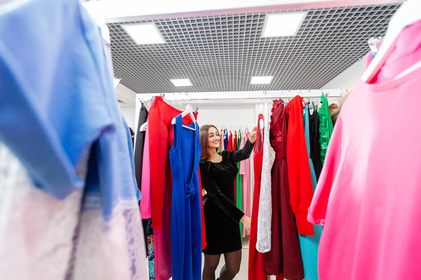 Счастливая молодая женщина выбирает одежду в торговом центре или магазине одежды. Продажа, мода, концепция потребительства
 - Фото, изображение