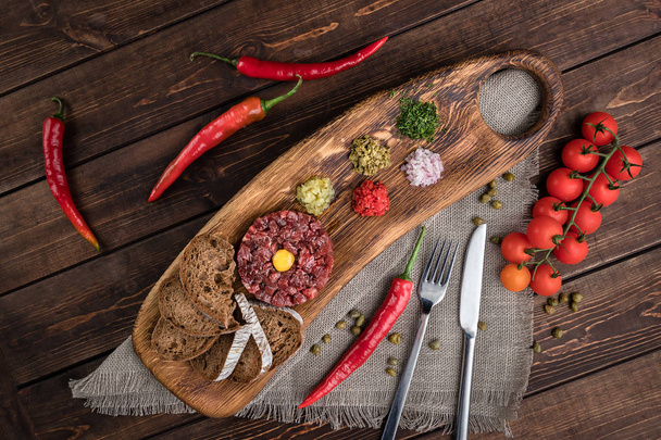 Top view on Beef Tartar with a raw egg yolk, rye bread and chilis on a wooden board and rough cloth with spoon and fork. Shot made from above on a dark rustic texture - Zdjęcie, obraz