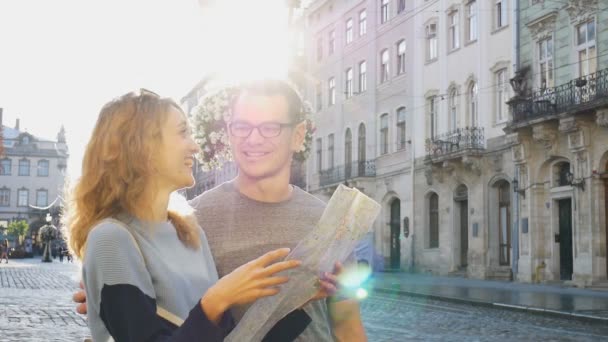 Happy young tourists couple holding a paper map of ancient European city early in the morning on empty square and pointing on some sightseeing place - Footage, Video