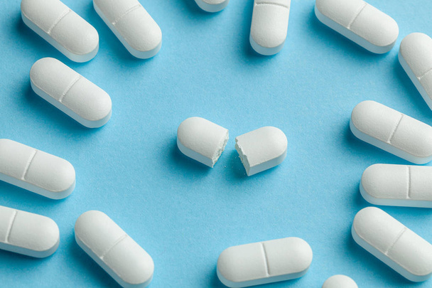 Valkoisia pillereitä sinisellä taustalla. Yksi tabletti on puolitettu, mikä pienentää lääkkeen annosta.
 - Valokuva, kuva
