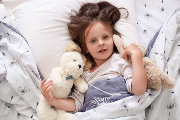 Horizontal shot of toddler girl in bed lying with his teddy bear at home, sleeppy child laying on linens with dandelion, looking directly at camera, has bcalm facial expression. Childhood concept. - Zdjęcie, obraz
