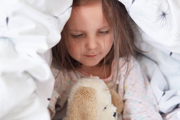 Indoor shot of adorable little child girl playing with teddy bear in bed under blanket, charming kid plays in morning before kinder garten, baby her teddy bear under covers looks at her white toy. - Foto, afbeelding