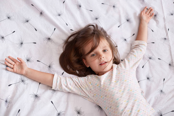 Top view of little girl laying in bed and spreading her arms on white cover with dandelion, charming kid with dark hair wearing pajama, looking at camera, relaxing after hard day. Children concept. - 写真・画像