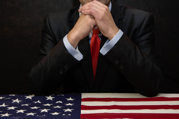 Ο πολιτικός-επιχειρηματίας σήκωσε τα χέρια του προσεκτικά καθώς καθόταν σε ένα τραπέζι καλυμμένο με αμερικάνικη σημαία, σκέψη και διάλογο. - Φωτογραφία, εικόνα