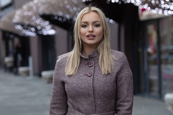 Blonde woman in brown woolen coat posing in city, modern building with festive lights on background. - Photo, image