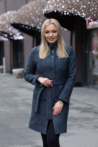 Portrait of young blonde woman in dark grey coat posing in city, modern building with festive shining lights on background. - Photo, Image