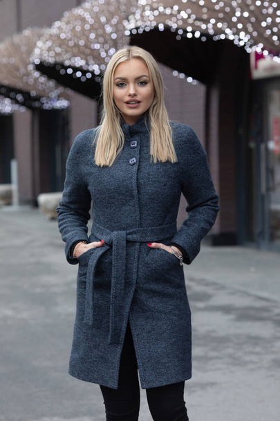 Portrait of young blonde woman in dark grey coat posing in city, modern building with festive shining lights on background. - Foto, imagen