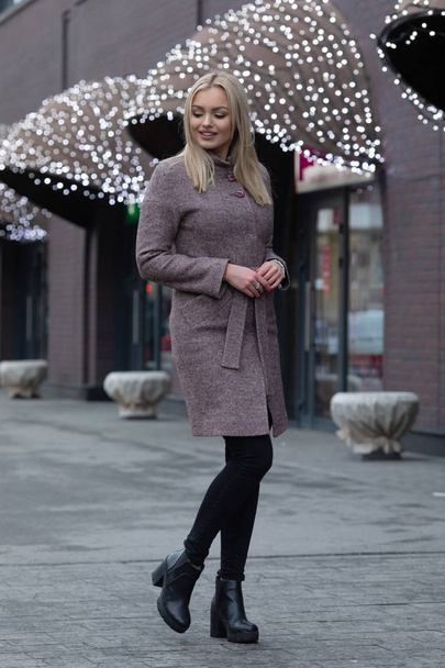Blonde woman in brown woolen coat posing in city, modern building with festive lights on background. - Fotografie, Obrázek