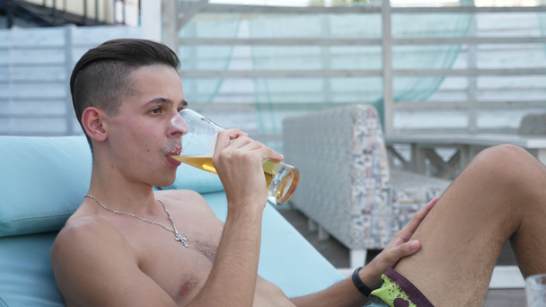 Jonge Brunet man liggend op een ligstoel en drinken bier op een zee Resort in de zomer spannend uitzicht op een jonge Brunet man zittend en liggend op een houten ligstoel geplaatst in een ruime parasol bedekt met een blauwe sluier. Hij drinkt bier en voelt zich gelukkig. - Video