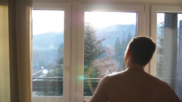 Otelin yatak odasında duran ve güneşli bir günde pencereden dışarı bakan tanınmayan atletik adam yakın. Yakışıklı genç adam sabahın erken saatlerinde güzel manzara zevk. Arka arka görünüm - Video, Çekim