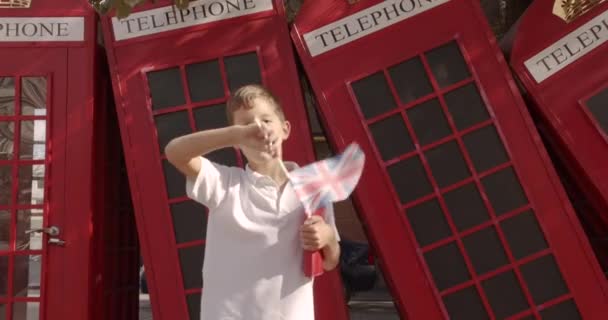 Lassú mozgás portréja Aranyos fiú integetett brit zászló áll a szabadban egyedül mosolyogva nézi kamera. A háttérben angol piros telefon fülkék. Utazás koncepció. - Felvétel, videó